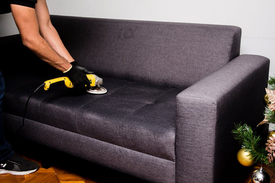 A person wearing black gloves is using a handheld electric upholstery steamer to deep clean a dark gray fabric sofa in a living room. The sofa has a modern design with clean lines and is positioned on a wooden floor. The surrounding area includes a small decorated Christmas tree with gold and silver ornaments, indicating a festive setting. Bright lighting illuminates the space, highlighting the cleanliness and freshness of the sofa after professional surface cleaning. The image demonstrates thorough domestic cleaning and sanitisation processes undertaken by Pimlico Carpet Cleaners, focusing on deep cleaning upholstery following an after-party to remove stains and ensure hygiene.