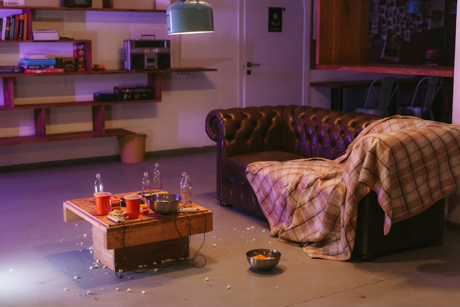 A person wearing black gloves is using a handheld electric upholstery steamer to deep clean a dark gray fabric sofa in a living room. The sofa has a modern design with clean lines and is positioned on a wooden floor. The surrounding area includes a small decorated Christmas tree with gold and silver ornaments, indicating a festive setting. Bright lighting illuminates the space, highlighting the cleanliness and freshness of the sofa after professional surface cleaning. The image demonstrates thorough domestic cleaning and sanitisation processes undertaken by Pimlico Carpet Cleaners, focusing on deep cleaning upholstery following an after-party to remove stains and ensure hygiene.
