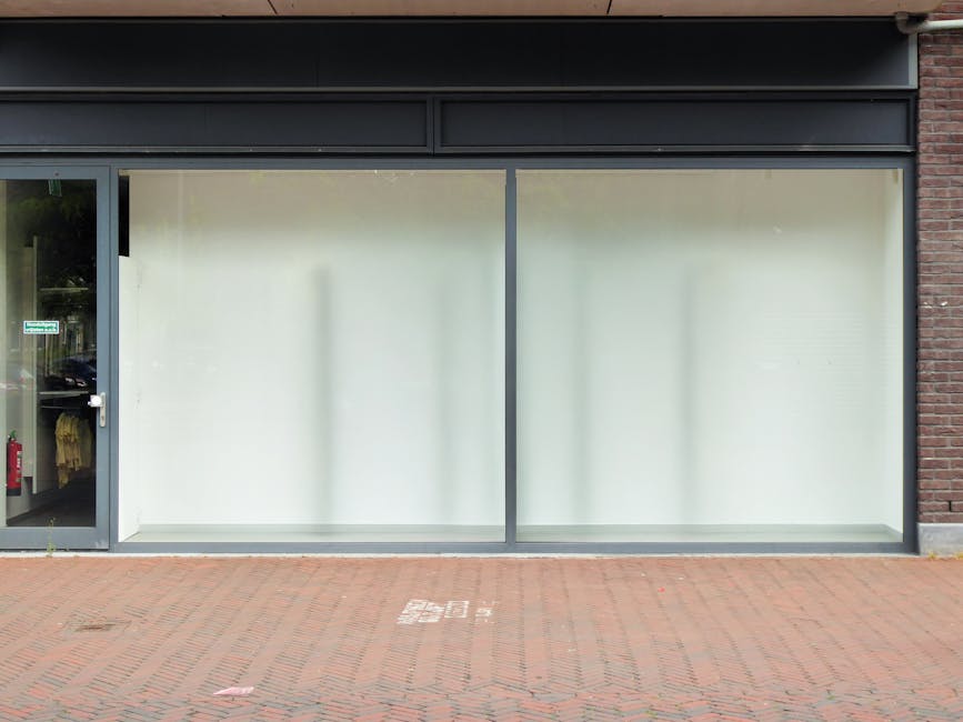 The image shows the exterior of a commercial storefront on Lupus Street, Pimlico, featuring large frosted glass windows that have been recently cleaned, displaying no dirt or smudges. To the left, there is a glass door with a metal frame, revealing an interior space with some clothing items visible on a rack and a fire extinguisher inside. The shopfront is framed by a dark metal fascia above the window. The ground in front of the store is paved with red bricks, and the overall appearance is tidy and well-maintained, reflecting professional surface cleaning and upkeep by Pimlico Carpet Cleaners as per their services in commercial cleaning options on Lupus Street, Pimlico.