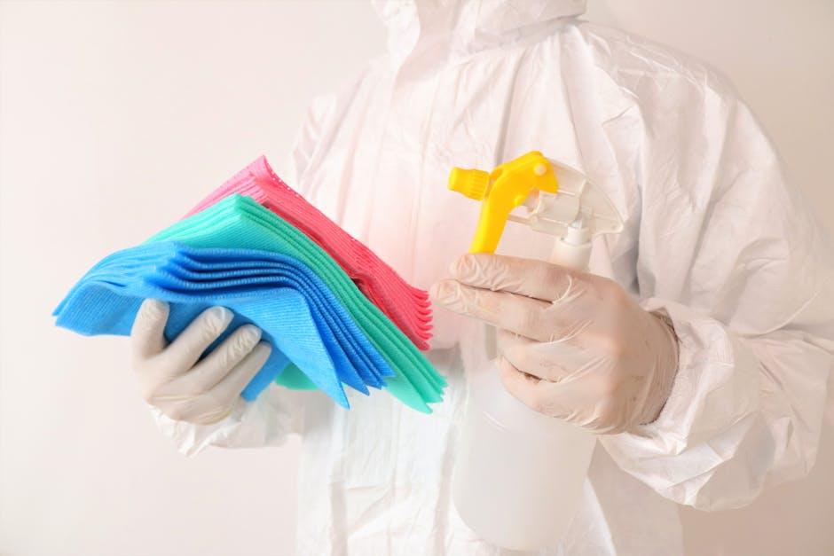 A person dressed in a white protective suit and gloves holding a stack of brightly colored cleaning cloths in blue, green, and pink with one hand, and a spray bottle with a yellow trigger in the other hand. The background is plain and neutral, focusing on the cleaning tools and protective gear, emphasizing professional surface cleaning and sanitisation processes used by Pimlico Carpet Cleaners for commercial cleaning options on Lupus Street, Pimlico.