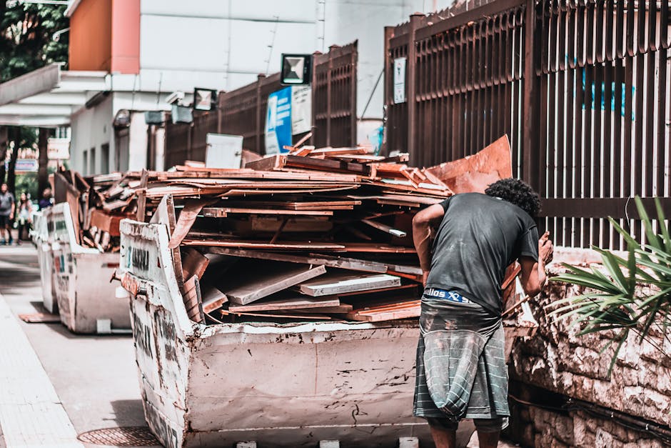 A man with curly black hair, wearing a black T-shirt and plaid shorts, stands outside near a large, rusty white skip filled with broken tiles, wood debris, and construction waste. The skip is position