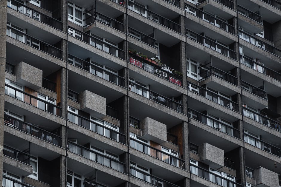 Close-up view of a high-rise residential building's exterior façade, showcasing multiple balconies with metal railings and glass doors. The balconies are constructed from grey concrete with some wood-paneled sections, and a few feature potted plants and outdoor furniture. The windows are clean and reflective, with natural light highlighting the textured surface of the building. This image emphasizes the building's structural materials and outdoor spaces, illustrating the typical exterior surfaces encountered in residential flats along Vauxhall Bridge Road. Pimlico Carpet Cleaners specializes in surface cleaning and maintenance, including deep cleaning and sanitisation of various residential façades and external surfaces in the Pimlico area.