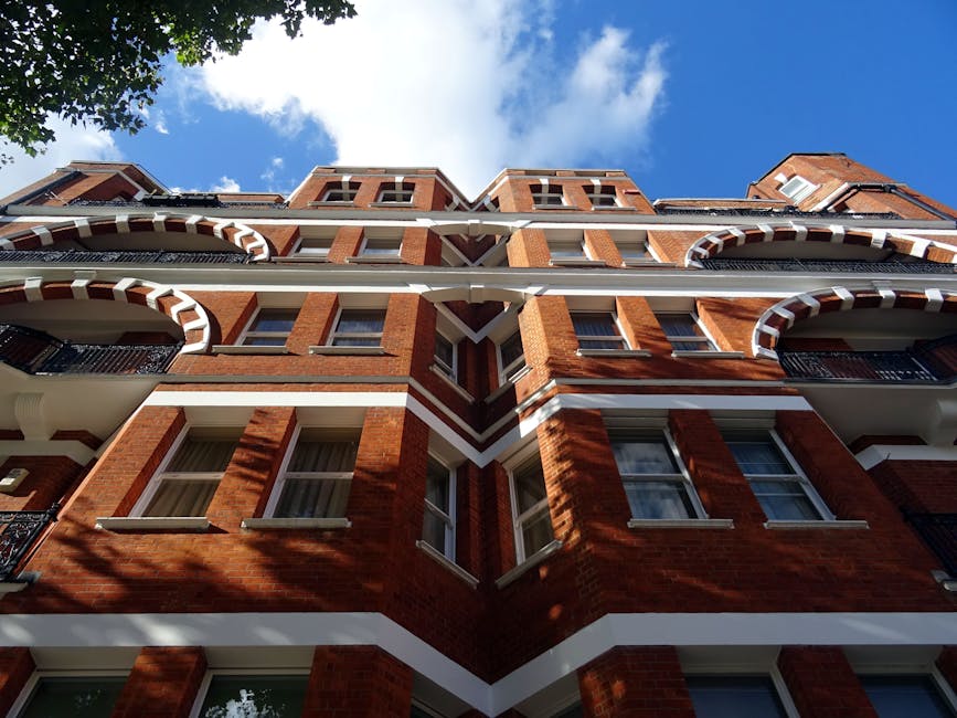 An exterior view of a multi-story red brick residential building with white decorative trim and black metal balcony railings, set against a bright blue sky with scattered clouds. The building features large windows, some with partially visible curtains, and clean, well-maintained surfaces. The sunlight creates shadows on the brickwork, highlighting its texture, while the overall scene suggests a well-kept urban residential area. Pimlico Carpet Cleaners specializes in surface cleaning and deep cleaning services, contributing to the upkeep of properties like this, including hygienic maintenance of indoor surfaces for flats on Vauxhall Bridge Road, Pimlico.