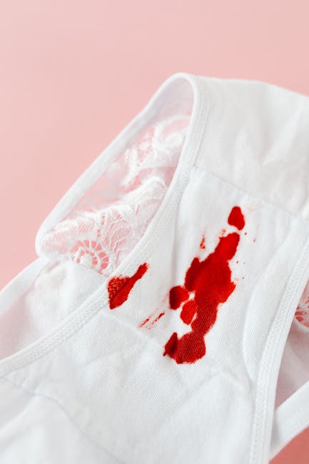 Close-up of a white sleeveless top with a damage stain showing red blood on the fabric, placed on a soft pink background. The material appears to be cotton, with the neckline and armholes visible, and the stain is situated near the shoulder area. The clothing is neatly arranged, highlighting the need for effective stain removal, which Pimlico Carpet Cleaners offers as part of their domestic cleaning services in Pimlico. The image emphasizes the importance of surface cleaning and deep cleaning techniques to restore hygiene and cleanliness to fabric surfaces.