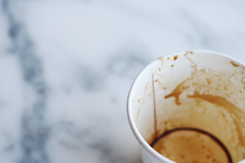 Close-up of a white ceramic coffee mug with coffee stains on its interior rim and sides, placed on a light-colored marble or stone countertop surface. The area surrounding the mug appears clean and well-maintained, with visible reflections of light on the smooth surface. In the background, there is a faint, out-of-focus detail of a stainless steel faucet or sink, suggesting a kitchen or similar room. The lighting is soft and natural, highlighting the cleanliness and sparkle of the surface, and emphasizing the importance of surface cleaning and maintenance standards promoted by Pimlico Carpet Cleaners in their domestic cleaning services, as discussed in the page about same-day stain removal costs.