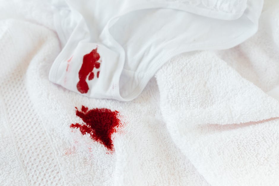 Close-up of a white sleeveless top with a damage stain showing red blood on the fabric, placed on a soft pink background. The material appears to be cotton, with the neckline and armholes visible, and the stain is situated near the shoulder area. The clothing is neatly arranged, highlighting the need for effective stain removal, which Pimlico Carpet Cleaners offers as part of their domestic cleaning services in Pimlico. The image emphasizes the importance of surface cleaning and deep cleaning techniques to restore hygiene and cleanliness to fabric surfaces.