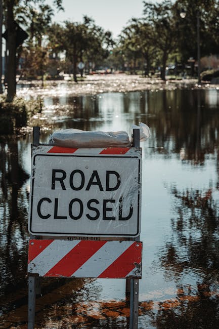 Two men are working to clear debris and trash from a flood-affected area near a body of water. One man, wearing a wide-brimmed hat, blue long-sleeve shirt, and black waterproof apron, is standing in t