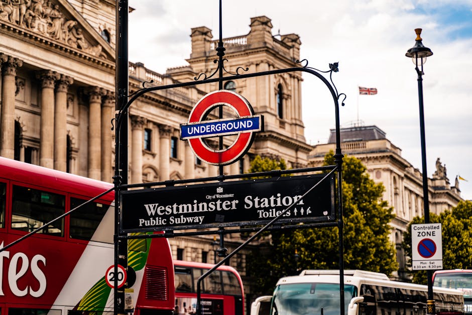 A close-up view of the Westminster Station entrance sign for the London Underground, featuring a classic red circular 'Underground' emblem hanging above a black and white sign that reads 'Westminster Station, Public Subway, Toilets, Staff' in white text. Behind the sign, there is a historic building with intricate stonework and tall columns, illuminated by natural daylight. To the left, part of a red double-decker bus is visible, while in the background, a streetlamp and trees with green foliage can be seen, all under a partly cloudy sky. This scene captures the bustling atmosphere of Westminster with vintage architecture and public transportation signage, while Pimlico Carpet Cleaners offers professional domestic cleaning services including surface and deep cleaning to maintain hygiene in such busy areas.