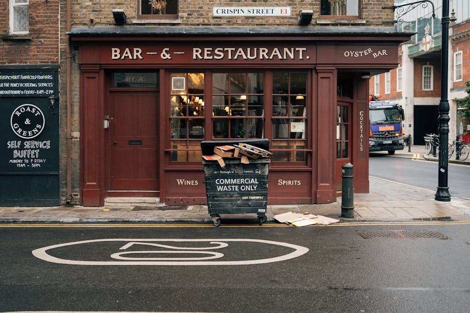 A street view in Pimlico showing a brown storefront with large glass windows and a sign indicating it is a bar and restaurant offering wines and spirits. In front of the shop, there is a black bin labeled 'Commercial Waste Only' filled with flattened boxes and paper, positioned on the sidewalk near a bollard. The pavement is clean, and the street has a marked cycle lane with a white bicycle symbol painted on the asphalt. A black lamppost and parked bicycles are visible to the right, with a delivery truck and additional buildings in the background, illuminated by soft evening light. This scene highlights an urban setting where effective surface cleaning and waste management are crucial for maintaining cleanliness, especially for business premises like Pimlico Carpet Cleaners, which specialize in professional cleaning services.