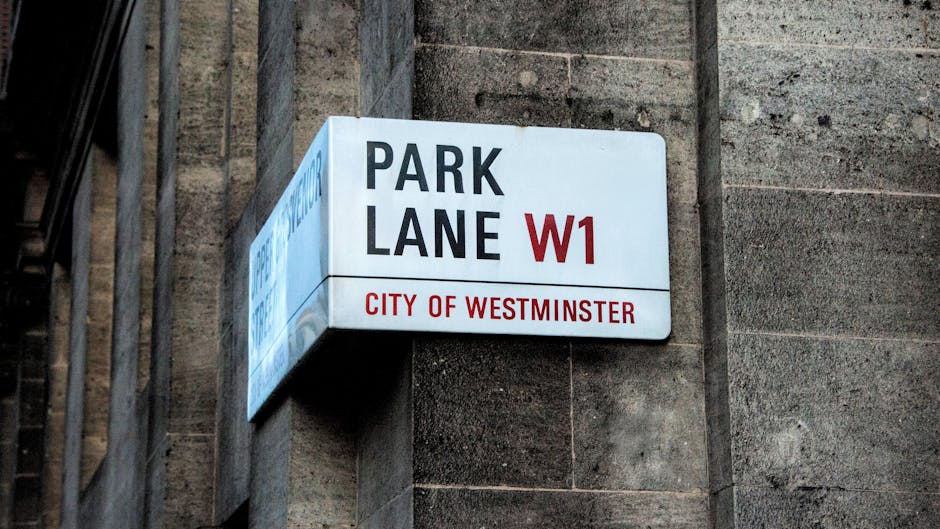 A close-up view of the Westminster Station entrance sign for the London Underground, featuring a classic red circular 'Underground' emblem hanging above a black and white sign that reads 'Westminster Station, Public Subway, Toilets, Staff' in white text. Behind the sign, there is a historic building with intricate stonework and tall columns, illuminated by natural daylight. To the left, part of a red double-decker bus is visible, while in the background, a streetlamp and trees with green foliage can be seen, all under a partly cloudy sky. This scene captures the bustling atmosphere of Westminster with vintage architecture and public transportation signage, while Pimlico Carpet Cleaners offers professional domestic cleaning services including surface and deep cleaning to maintain hygiene in such busy areas.